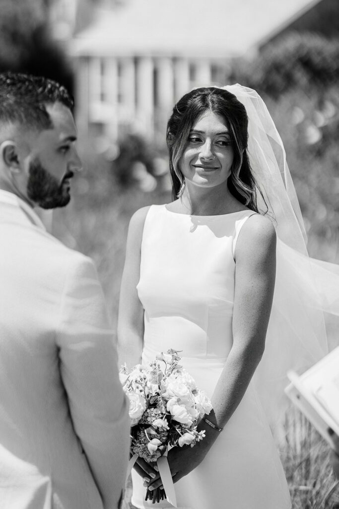 A bride in a boatneck sleeveless dress is standing opposite the groom holding her bouquet at hip level and smiling off to the side. 