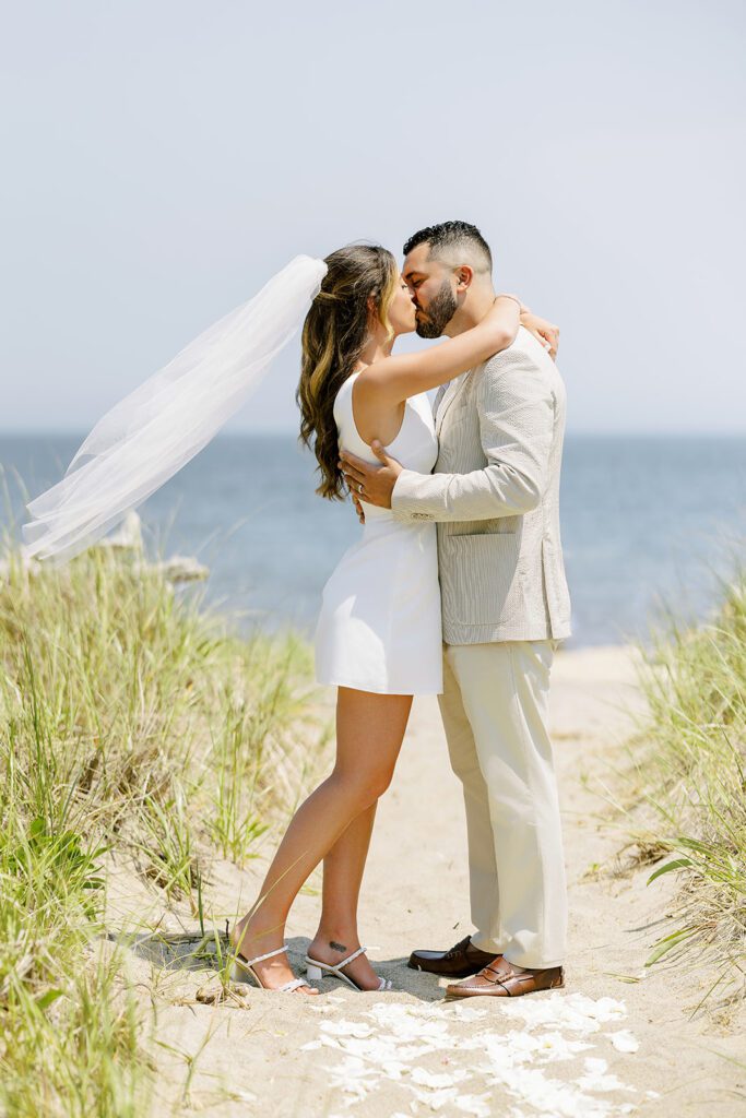 Brunette bride and brunette, bearded groom embrace in a kiss in the dunes of a beach. Her veil is flowing out behind her and her arms are wrapped around his neck. 