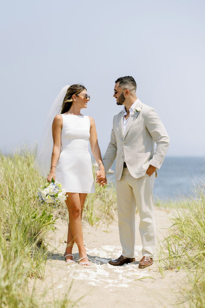 A bride in a white, sleeveless mini-dress wearing a mid-length veil is holding a bouquet of flowers in one hand and with her other, holding the hand of her brunette and bearded husband in a neutral suit. They are standing on a sandy path between the dunes with the ocean visible in the background. White petals are scattered beneath their feet on the sand. 