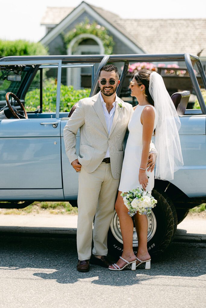 A brunette, bearded groom wearing sunglasses and has his arm around his brunette bride who is wearing a white, sleeveless mini dress and holding a bouquet down by her side. She is smiling at him while he is smiling straight ahead. They are standing in front of a blue convertible SUV. 