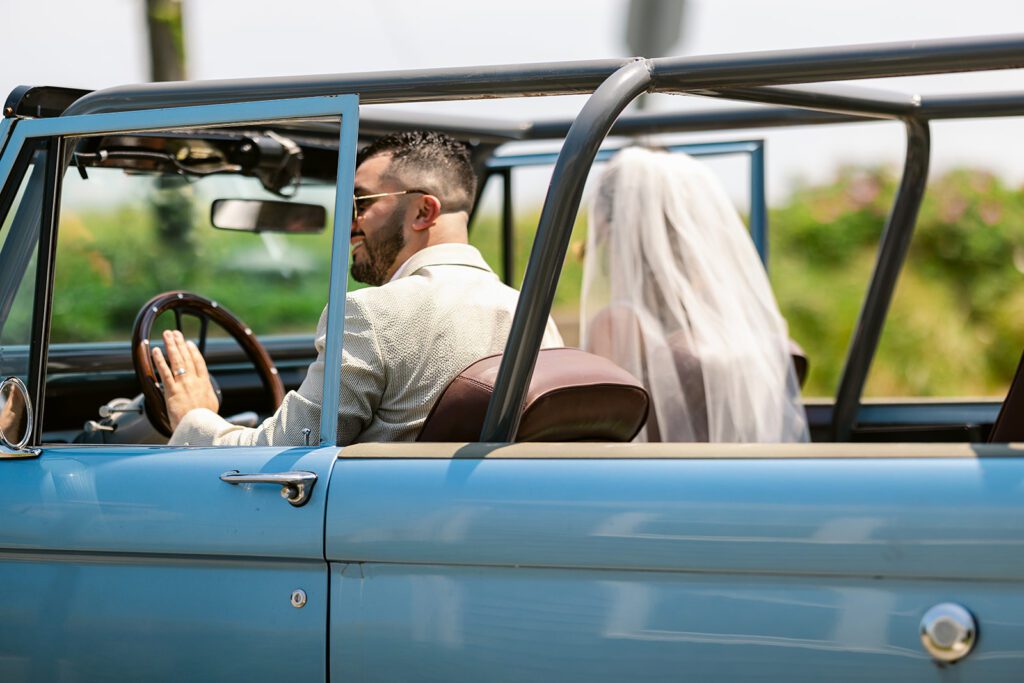 A bride and groom are sitting inside a blue convertible SUV - the groom is wearing sunglasses with his hand on the wheel while the bride's veil is flowing over the back of the front seat. 