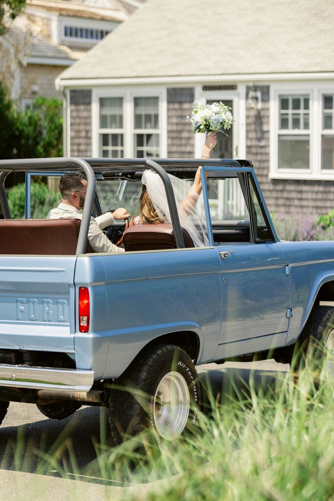 A bride and groom are sitting inside a blue Ford bronco convertible - the bride is holding her bouquet up in the air above the front windshield. 