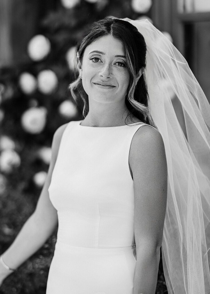 A bride in a boatneck, sleeveless dress is smiling with her veil looking very full behind her. 