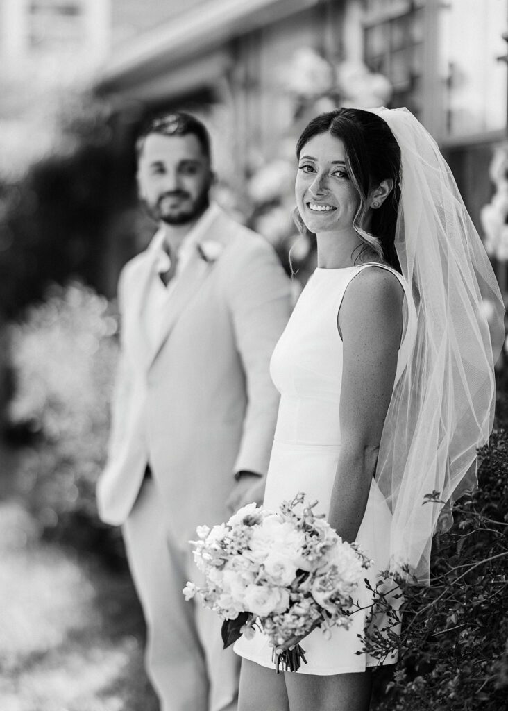 A bride wearing a veil and holding a bouquet is handing the hand of her groom who is standing off to the side/behind her and she is smiling. He is looking at her and smiling. 