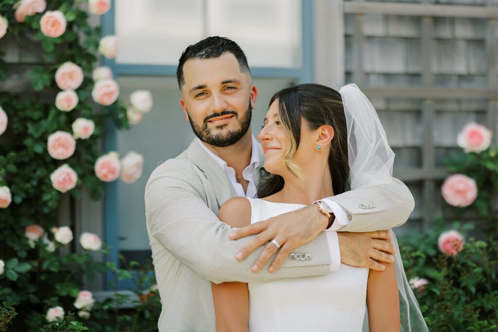 A brunette, bearded groom is smiling straight ahead with his arms around his brunette bride's shoulders. She is smiling up at him wearing a white sleeveless dress and white veil. 