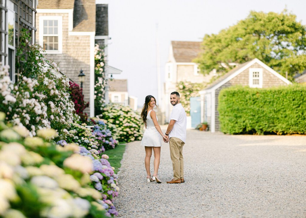 A brunette woman a white mini dress is holding hands with a brunette bearded man who is wearing a white short sleeve shirt and khakis. They are walking on a shell path surrounded by flowering bushes and green hedges and are looking behind them smiling. 