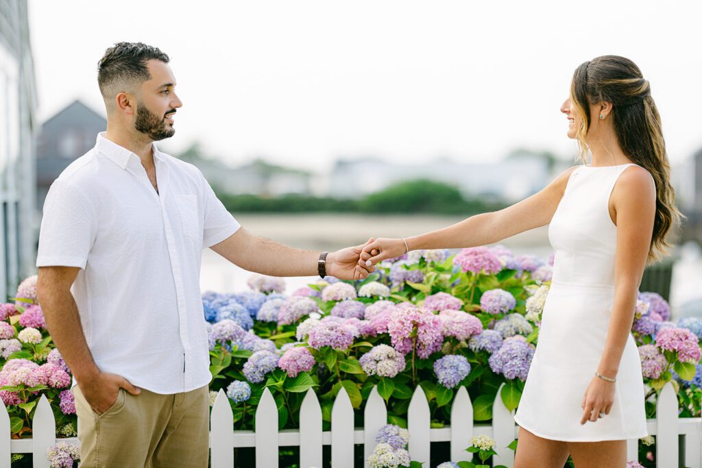 A brunette, bearded man in a white button down and khaki's is holding the hand of a brunette woman in a white, sleeveless mini dress. They are facing each other with their arms outstretched in front of a white picket fence with hydrangeas behind it. 