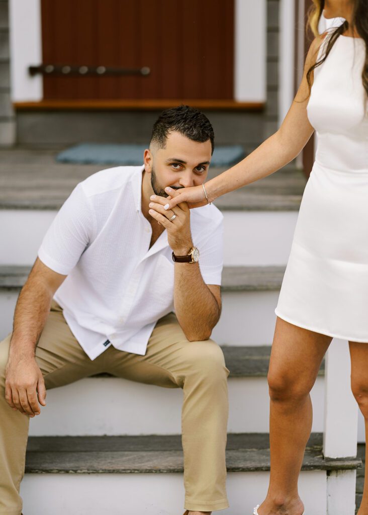 A brunette, bearded man is sitting on some steps and kissing the hand of a woman standing in front of him in a white mini dress. 