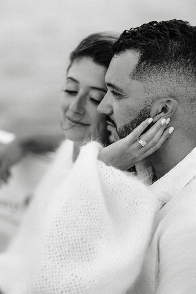 A woman is leaning her head into a man's head and smiling with a downward gaze- she has her hand cupping his face and he is staring straight ahead. 