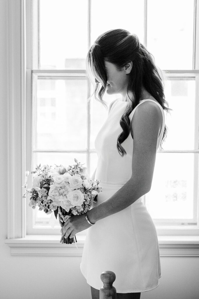 A bride is standing in front of a window in a sleeveless mini dress holding a bouquet of flowers. Her long hair is cascading over her shoulders with delicate tendrils hiding her face. 