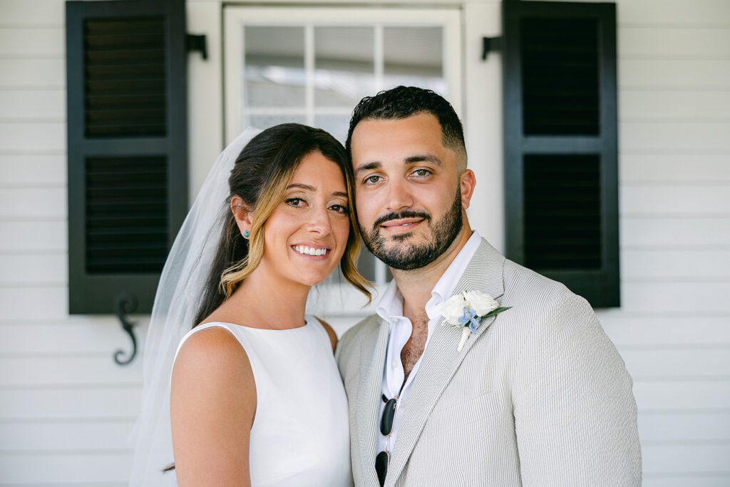 A brunette bride and a brunette, bearded groom are standing side by side with their temples touching and smiling. 