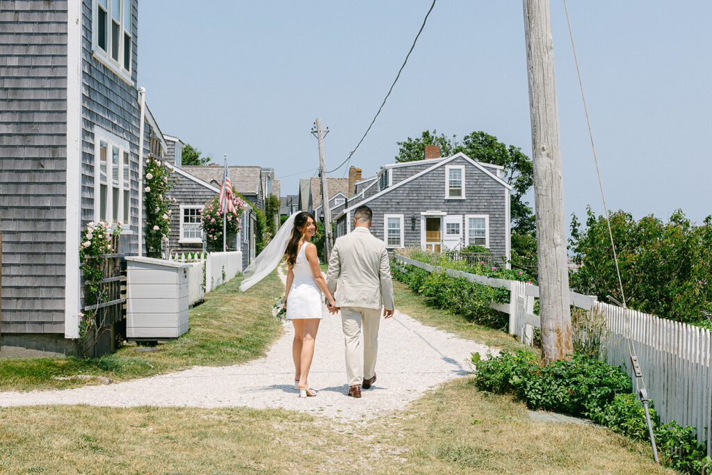 A bride and groom are walking side-by-side, hand-in-hand, down a shell pathway. She is looking back and smiling with her veil blowing in the wind. 