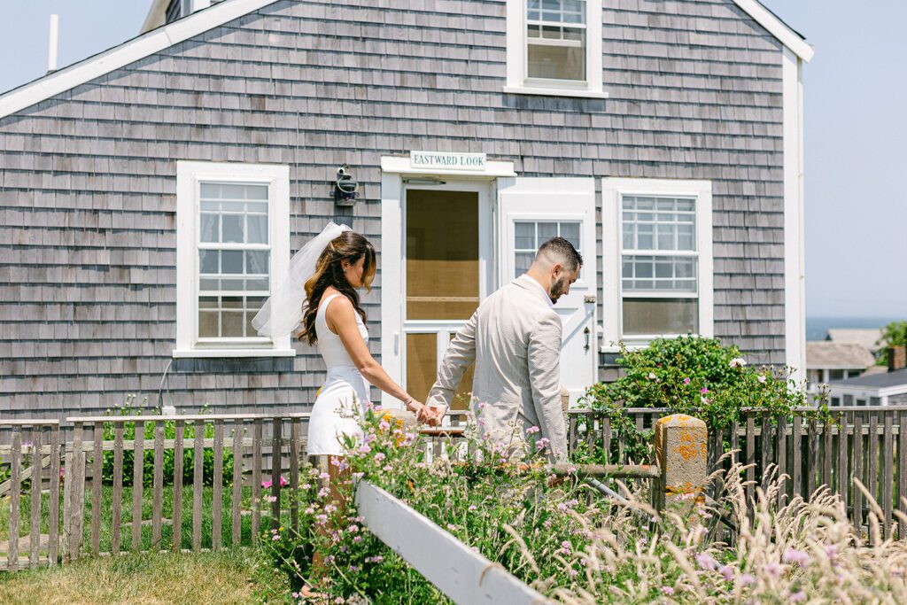 A bride and groom are walking hand-in-hand, he is leading her down a grassy walkway in front of a clapboard cottage. 