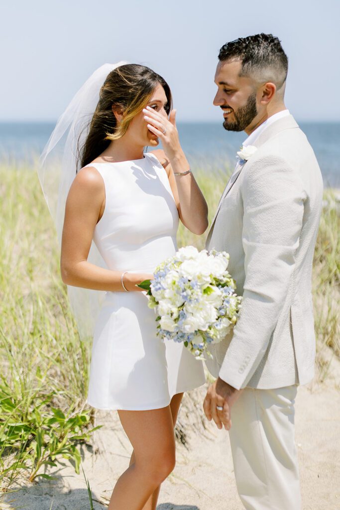 A brunette bride in a white boatneck mini dress is wiping away a tear. The brunette, bearded groom is standing in front of her in a neutral suit and smiling. 