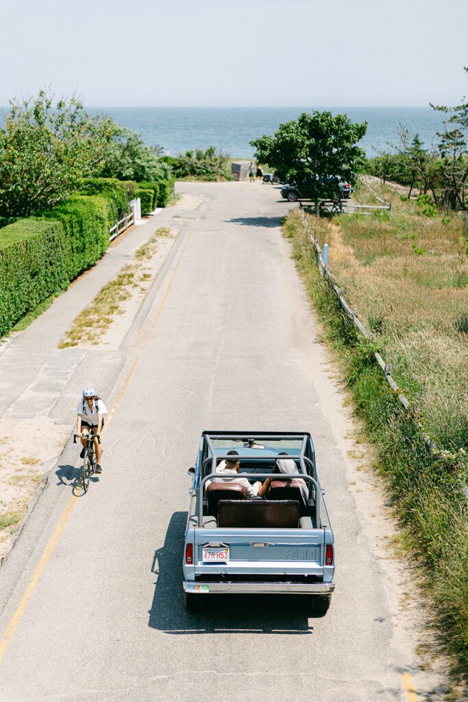 An aerial view of a bride and groom driving down a road towards the ocean in a blue Ford Bronco convertible. 