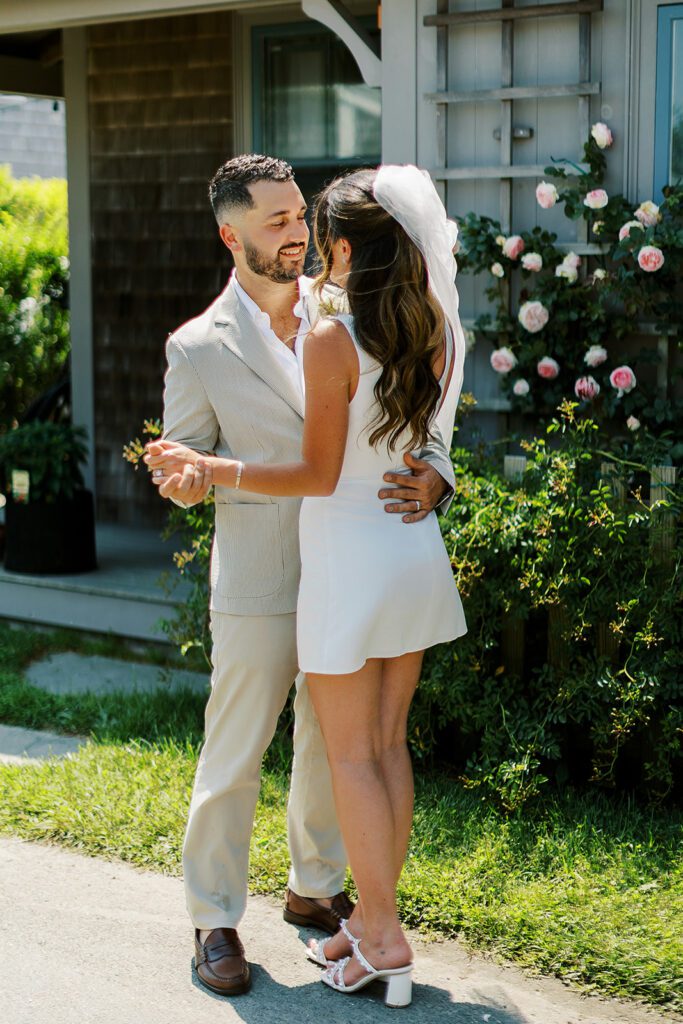 A brunette and bearded groom is holding his brunette bride wearing a white mini dress closely and they are mid-dance. 