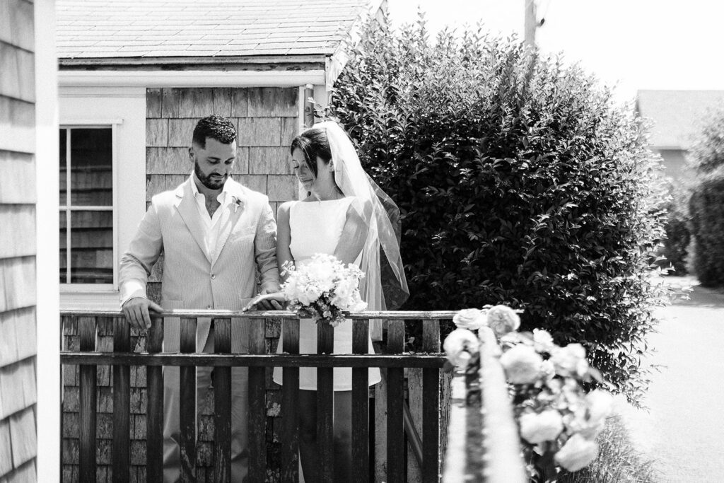 Bride and groom are standing behind a picket fence shoulder to shoulder. She is holding out her left hand and they are both looking down at it. 