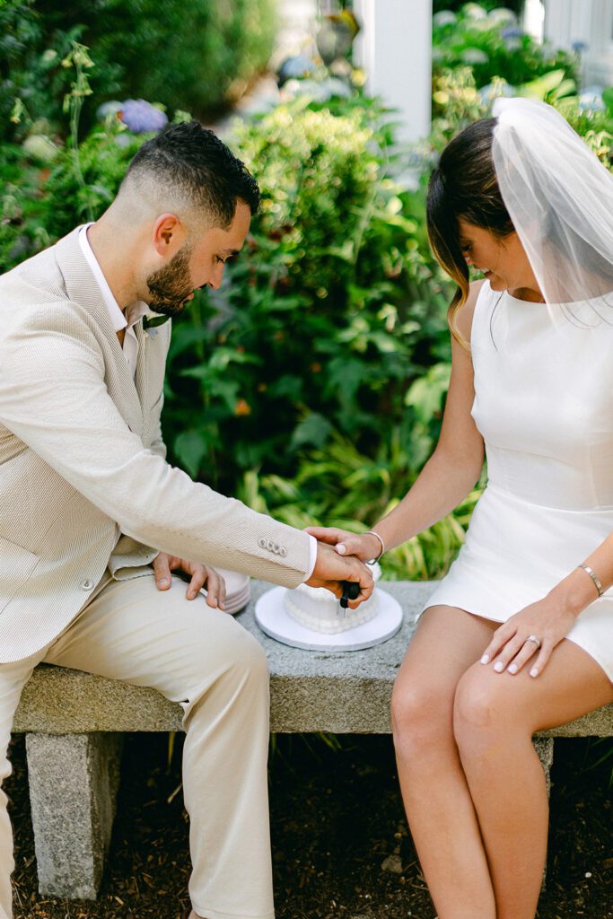 A bride and her groom are sitting on a stone bench. He is getting ready to cut a cake and her hand is resting on top of his hand as he does so. 