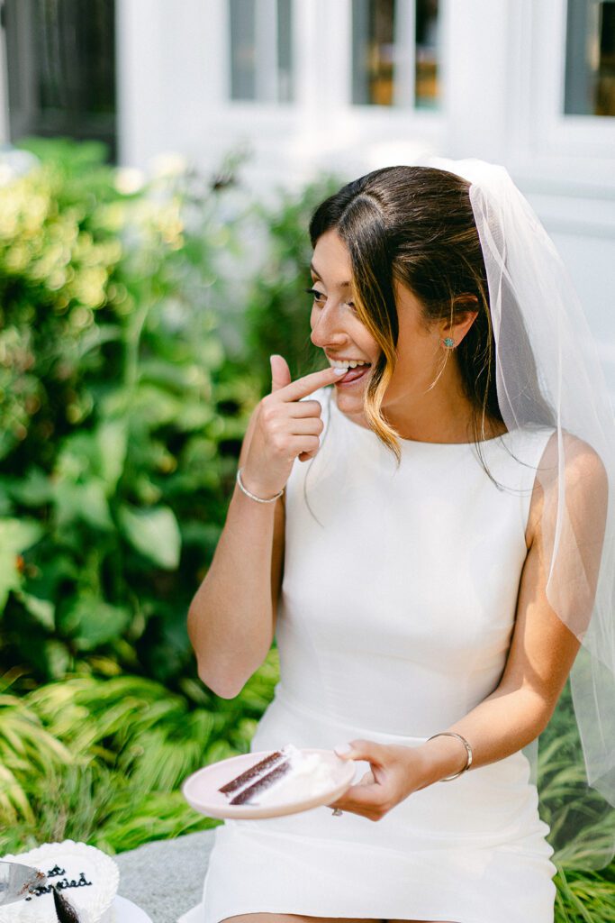 A brunette bride in a white, boatneck, sleeveless dress wearing a veil is holding a plate with a piece of cake in one hand and licking her finger on the other hand while smiling. 