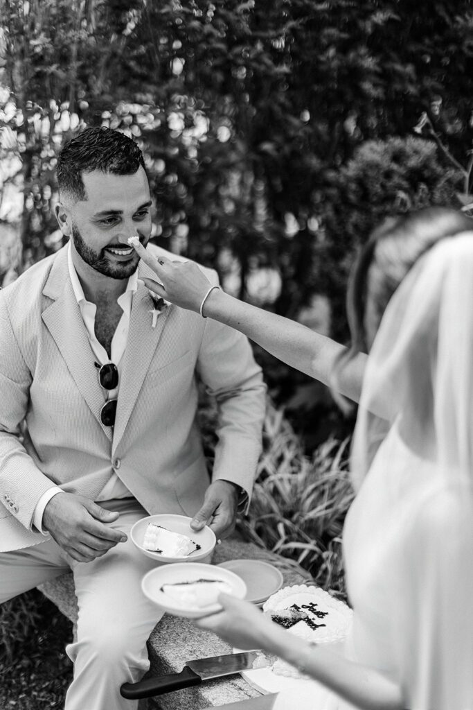 A bride is putting a putting a swipe of frosting on her grooms nose and he is smiling at her. Both are holding plates with a slice of cake on each while sitting on a bench. 