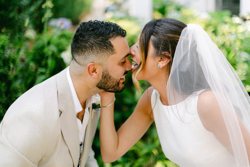 A brunette bride in a white tulle veil and while sleeveless dress is licking the frosting off of her smiling brunette and beaded groom's nose. 