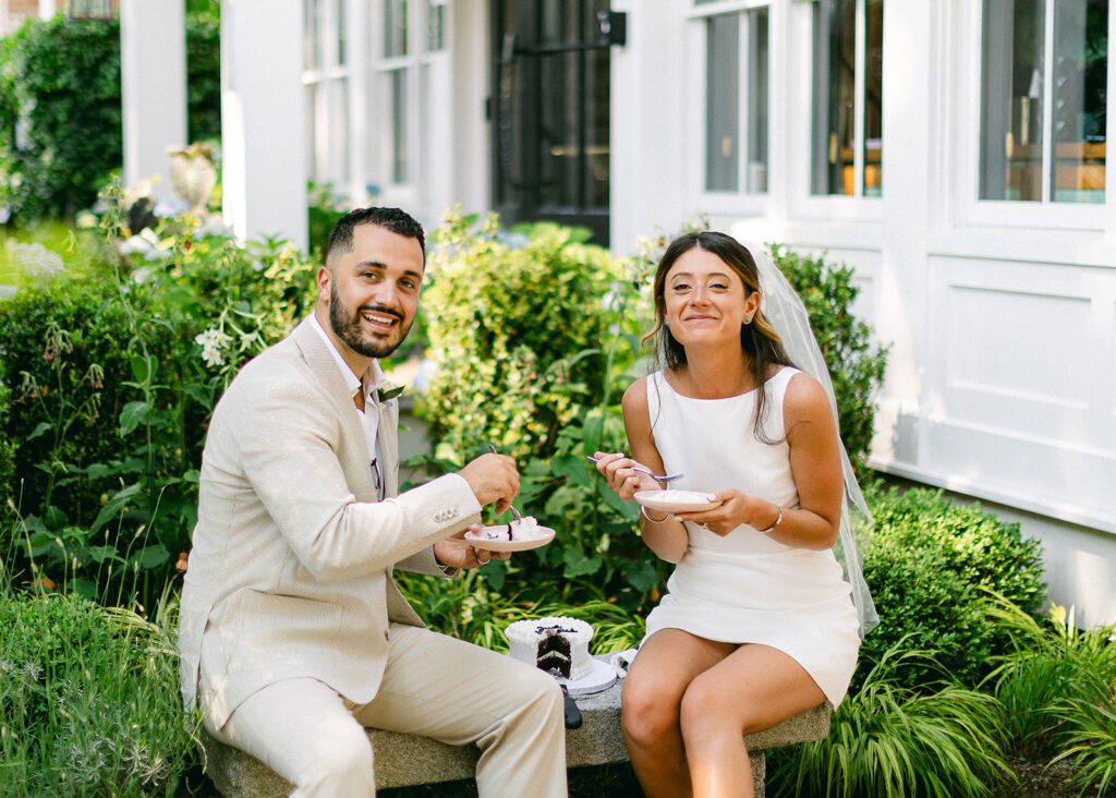 A bride and groom are sitting on a stone bench eating their wedding cake and smiling. 