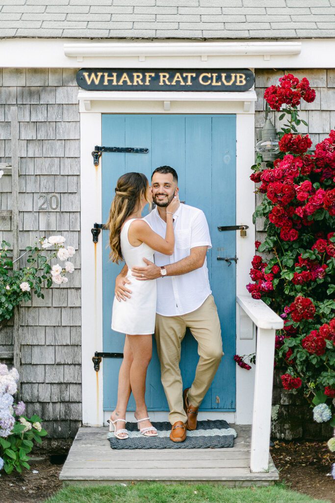 A brunette woman in a white min dress is standing next to a brunette, bearded man and kissing his cheek. He has his arms around her waist and is smiling. They are standing in front of a blue door of a clapboard house with red climbing roses on one side of them. 