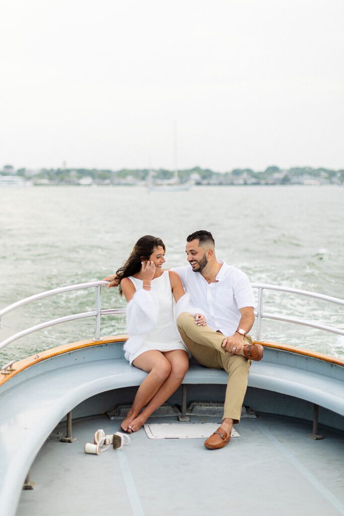 A brunette woman in a white sleeveless dress is sitting next to a brunette, bearded man on a boat.She has one arm resting on his leg, the other pulling hair out of her face, and is smiling at him. He is smiling at her with one arm around her. The water and land is visible behind them. 