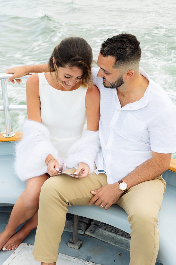 A brunette woman is snuggled in next to a brunette, bearded man on a bench on a boat. She is looking at something she is holding in her hands and smiling, and he has his arm around her and is smiling at her. 