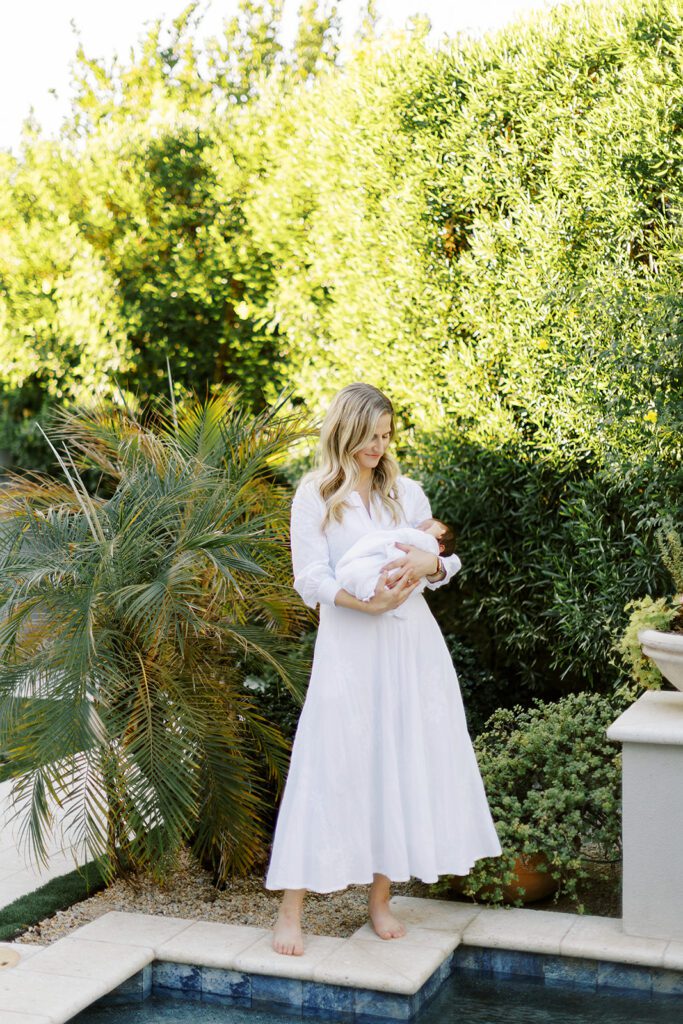 Blonde mom is standing on the edge of a pool in front of lush greens in a long-sleeved, white maxi dress holding her dark-haired newborn baby in her arms all swaddled in a white muslim blanket. 