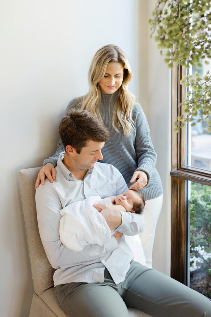 Brunette dad in a button-down shirt is siting and smiling down at his newborn baby who is swaddled in a white blanket and sleeping in his arms. Blonde mom in a gray sweater and white jeans is standing behind him and resting one hand on the baby's head and one hand on her husband's shoulder. 