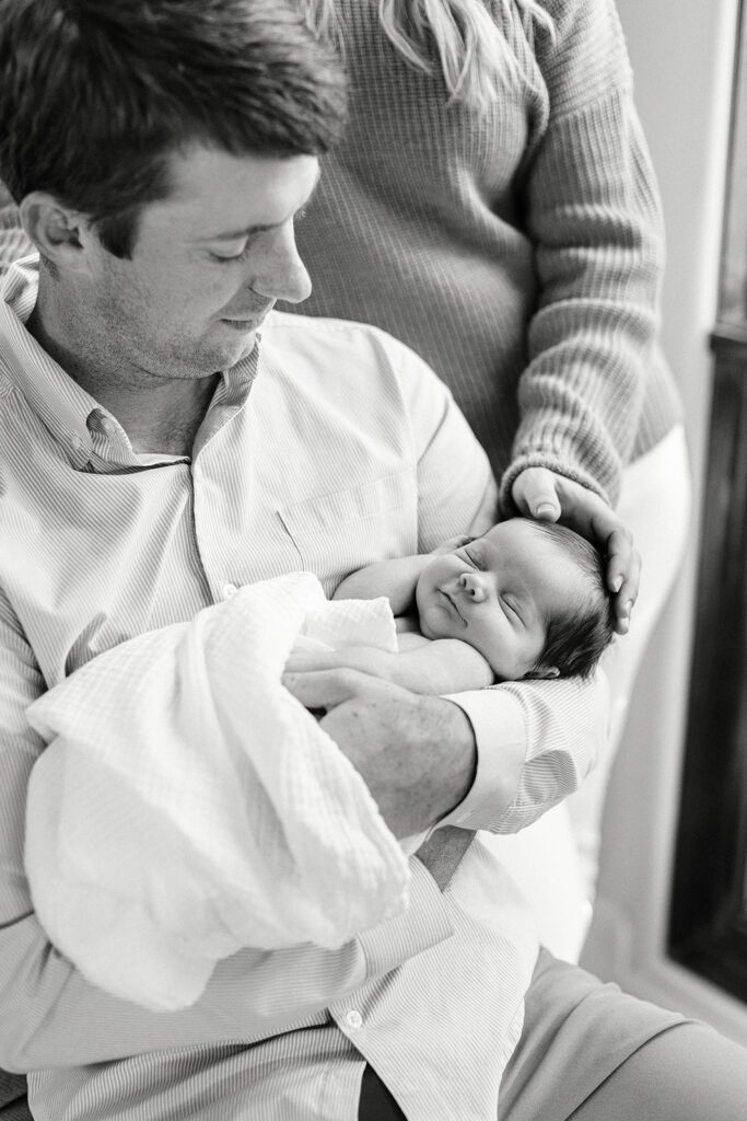 Dad is siting and smiling down at his newborn baby who is swaddled and sleeping in his arms. Mom i standing behind him and resting a hand on the baby's head. 