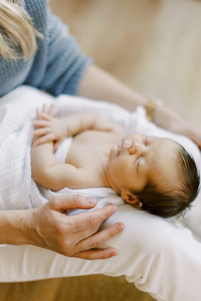 Dark-haired newborn baby is sleeping & swaddled in a white muslim blanket and laying on mom's legs with hands resting belly. 