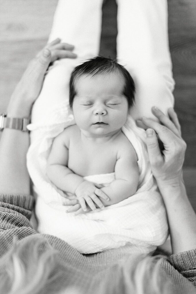 Dark-haired newborn baby is sleeping & swaddled in a white muslim blanket and laying on mom's legs with hands resting belly. 