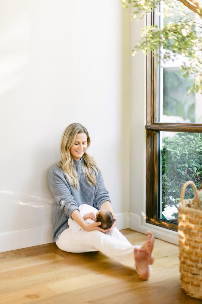 Blonde mom in a grey sweater and white jeans is sitting on the floor against the wall with legs outstretched - her newborn baby is resting on her legs and she is cupping the dark-haired head with both hands and smiling down at baby. 