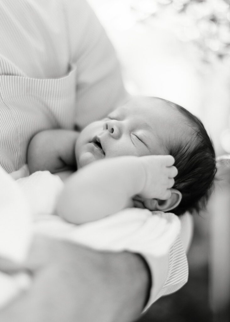 Dark-haired newborn baby is sleeping in the nook of an adult's arms with both hands resting on each cheek. 