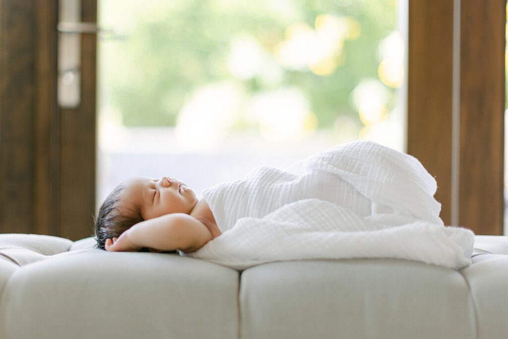 Dark-haired newborn baby is naked and swaddled in a white blanket, sleeping on an upholstered, tufted bench with both arms raised and resting on either side of the head. 