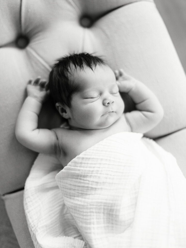 Dark-haired newborn baby is naked and swaddled in a white blanket, sleeping on an upholstered, tufted bench with both arms raised and resting on either side of the head. 