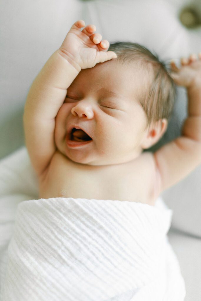 Dark-haired newborn baby is swaddled in a white muslim blanket and is mid-stretch with arms raised, eyes closed, and mouth slightly agape with the tongue visible against the gums. 