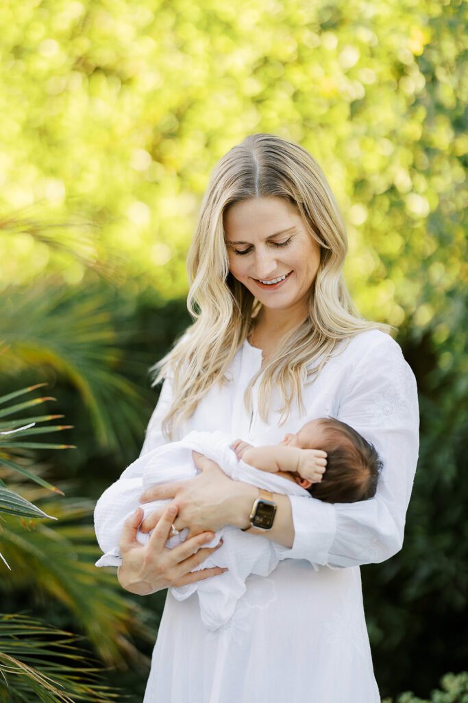 A blonde, smiling mom is standing in front of lush greens in a long-sleeved, white dress holding her dark-haired newborn baby in her arms all swaddled in a white muslim blanket. 