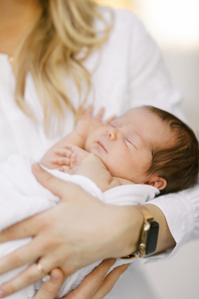 A dark-haired newborn baby is snuggled up in a white muslim blanket and sleeping in mom's arms up against her chest. 