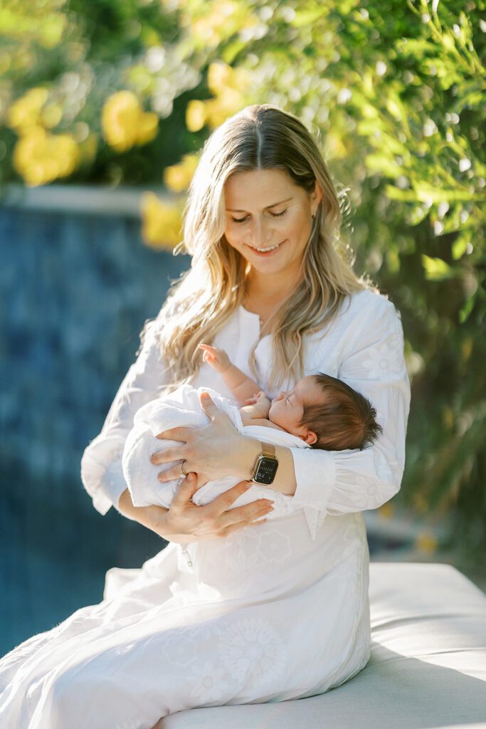 Blonde haired mom in a white long-sleeve maxi dress is sitting on a bench in front of lush greens outside and smiling down at her swaddled dark-hair newborn baby who is sleeping in her arms. 