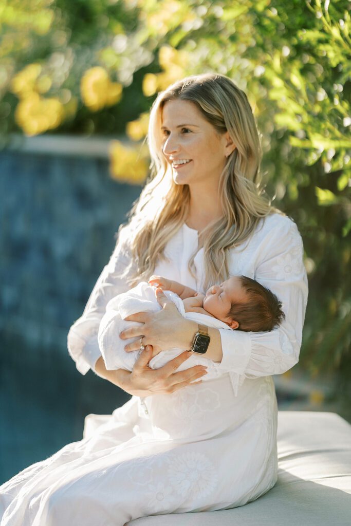 Blonde haired mom in a white long-sleeve maxi dress is sitting on a bench in front of lush greens outside and smiling off into the distance while her swaddled dark-hair newborn baby is sleeping in her arms. 