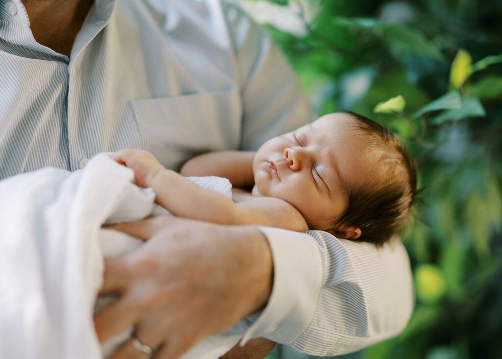 Dark-haired newborn baby is sleeping sounding in the nook of an arm with one hand tucked beside the head, swaddled in a white muslim blanket. 