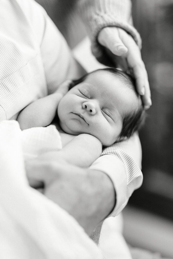 Dark-haired newborn baby is sleeping sounding in the nook of an arm with one hand tucked beside the head. The hand of another person is gently resting on the baby's head. 