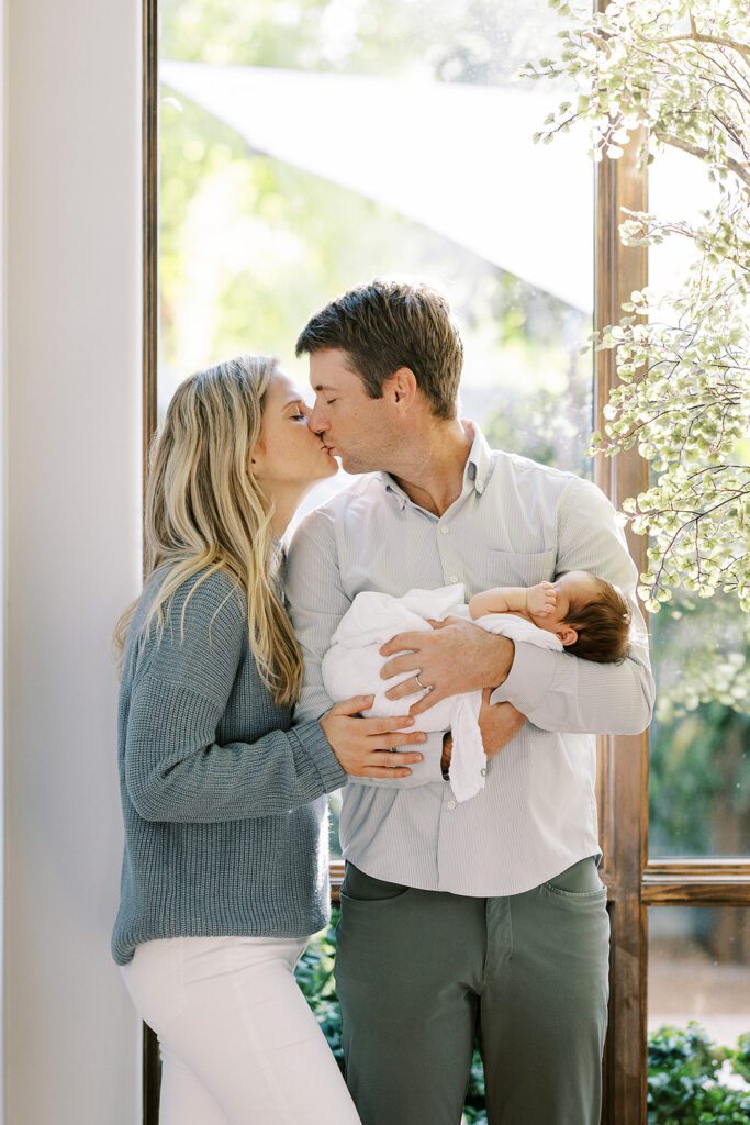 Brunette dad is wearing a button-down shirt and gray slacks while holding a dark-hared newborn baby swaddled in a white muslim blanket while kissing his blonde haired wife standing beside him who herself is wearing a grey sweater and white jeans. 