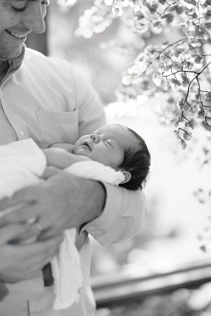 Dad is standing by an indoor tree with small fan-shaped leaves, holding dark-haired newborn, sleeping, swaddled baby in his arms and smiling down at baby. 