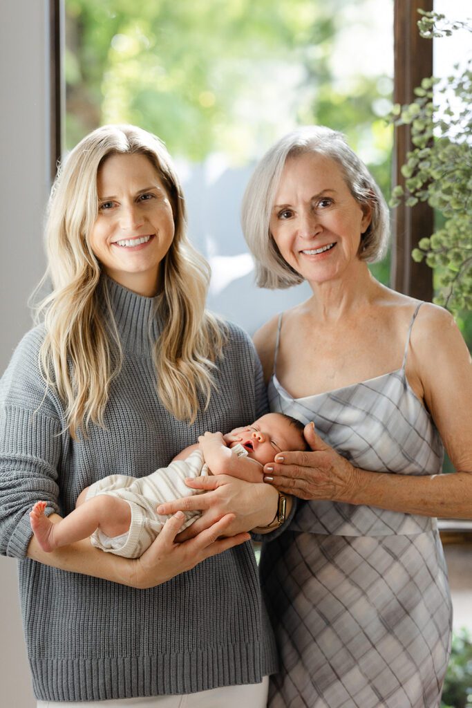  Blonde mom is wearing a grey turtleneck sweater, smiling, and holding her newborn baby. Grandma, with a short, grey bob & wearing a gray plaid slip dress, is standing right beside her resting one hand on the baby's cheek, also smiling. 