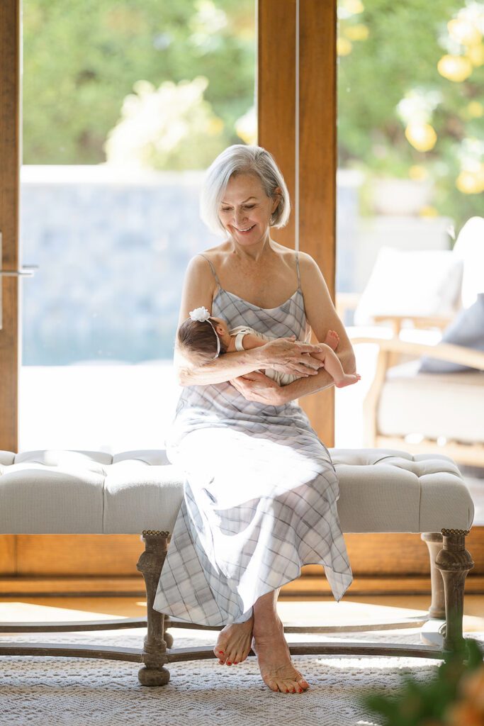 A woman wearing a grey, plaid slip dress donning a short grey bob is sitting on a tufted upholstered bench and holding a dark-haired baby girl wearing a little headband with a white flower in her arms. 