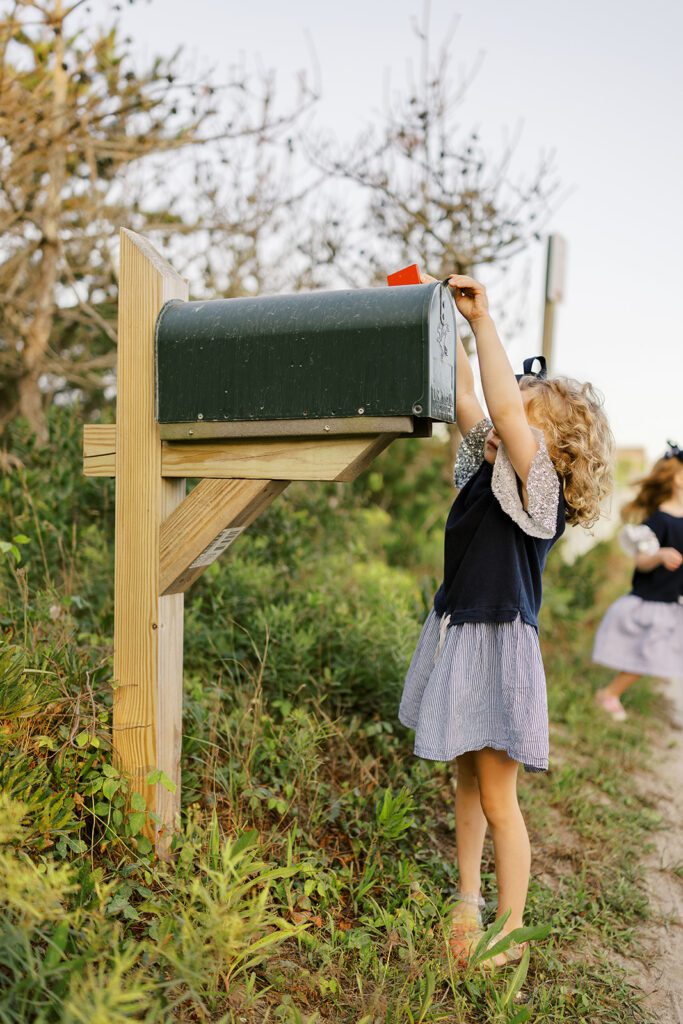 A little girl is reaching up to close her mailbox and put the red flag up. Her face is hidden behind her arm but light brown curls are cover her neck. 